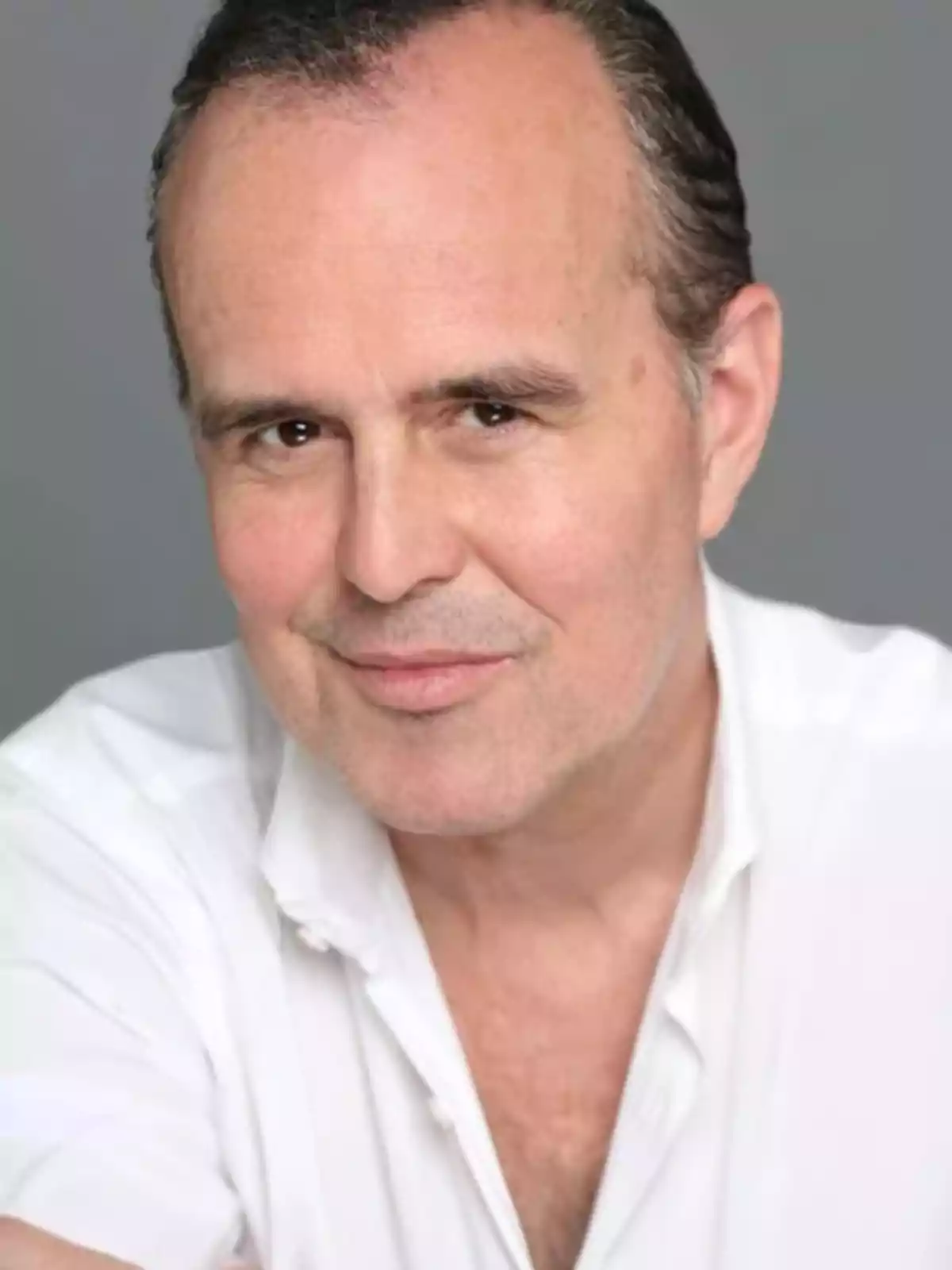 Hombre de mediana edad con cabello corto y oscuro, vestido con camisa blanca y fondo gris, mirando a la cámara con una expresión amigable Hombre de mediana edad con cabello corto y oscuro, vestido con camisa blanca y fondo gris, mirando a la cámara con una expresión amigable