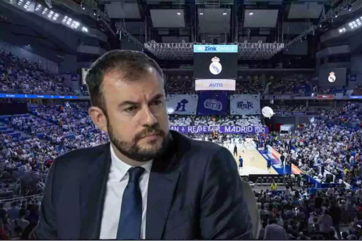 Hombre con barba y traje oscuro en primer plano con un estadio de baloncesto lleno de aficionados al fondo