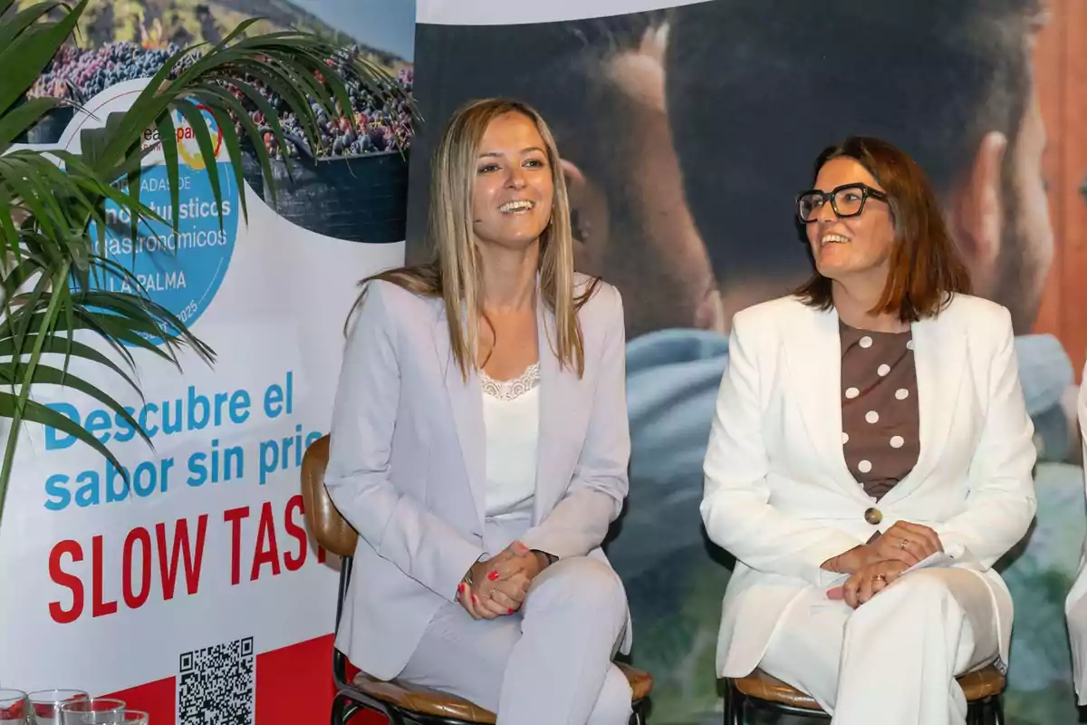Dos mujeres sentadas sonríen durante un evento promocional con un cartel de fondo que dice Descubre el sabor sin prisa SLOW TASTE