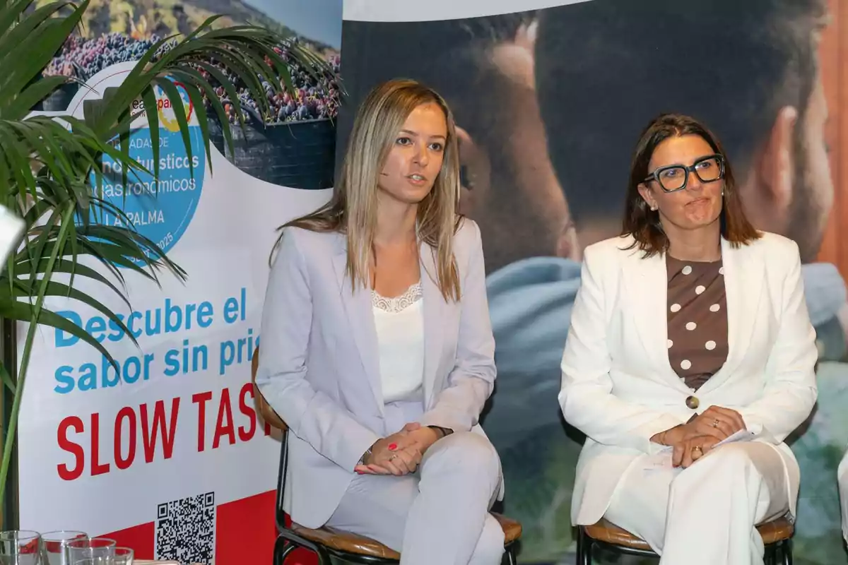 Dos mujeres sentadas en sillas durante un evento, ambas vestidas con trajes claros, una de ellas lleva gafas grandes y fondo con carteles promocionales y plantas.