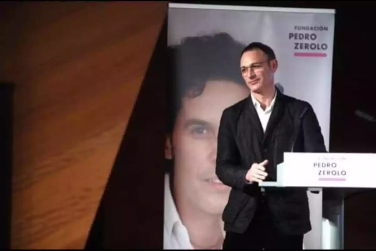 Hombre de pie junto a un atril durante un evento de la Fundación Pedro Zerolo con un cartel de fondo que muestra un rostro masculino Hombre de pie junto a un atril durante un evento de la Fundación Pedro Zerolo con un cartel de fondo que muestra un rostro masculino