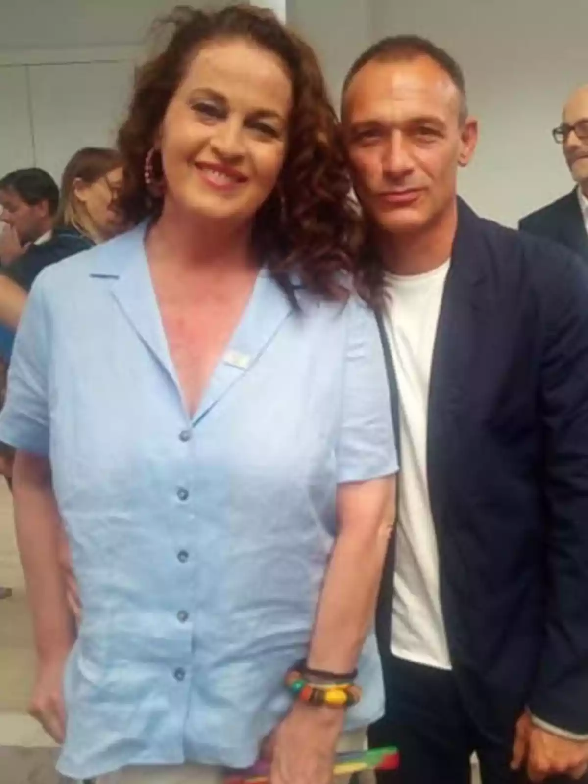 Dos personas posan juntas y sonríen en un ambiente interior con más gente al fondo Dos personas posan juntas y sonríen en un ambiente interior con más gente al fondo