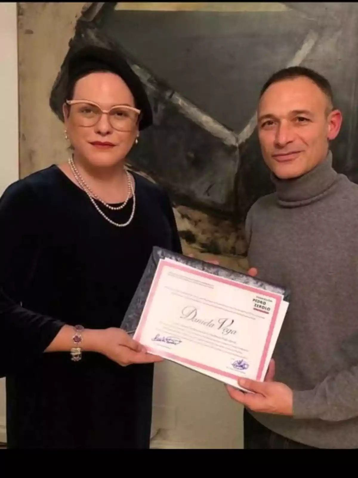 Dos personas posan juntas mientras sostienen un diploma enmarcado frente a una pintura abstracta. Dos personas posan juntas mientras sostienen un diploma enmarcado frente a una pintura abstracta.
