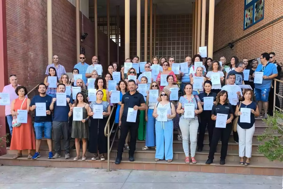 Un grupo numeroso de personas está de pie en unas escaleras frente a un edificio sosteniendo hojas blancas con mensajes impresos en señal de apoyo o protesta Un grupo numeroso de personas está de pie en unas escaleras frente a un edificio sosteniendo hojas blancas con mensajes impresos en señal de apoyo o protesta