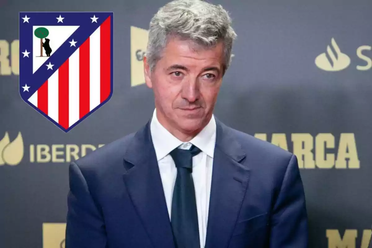 Hombre de cabello canoso con traje azul y corbata, de pie frente a un fondo con logotipos y el escudo del Atlético de Madrid a su lado izquierdo.