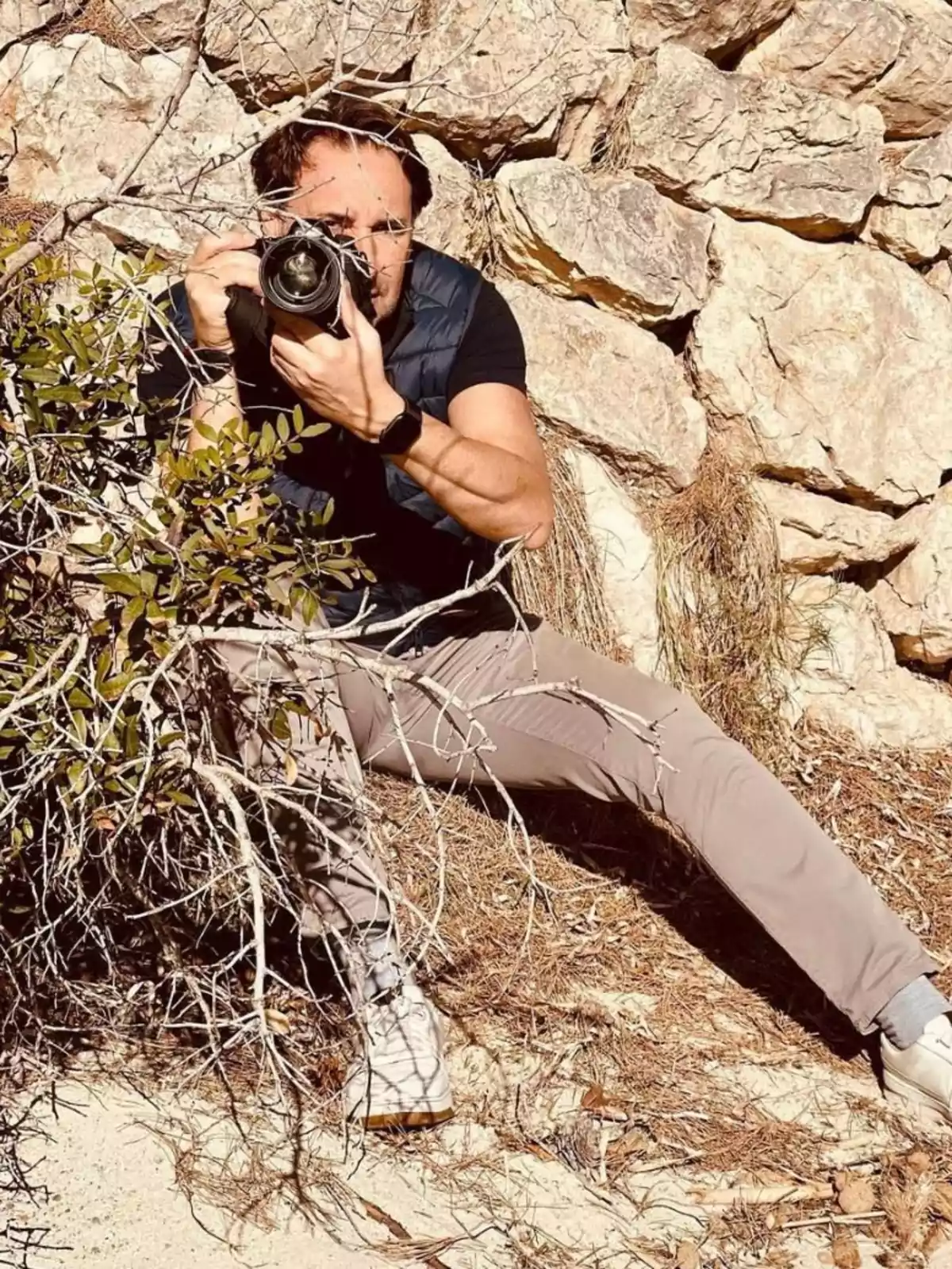 Hombre agachado entre ramas y rocas tomando una fotografía con una cámara profesional Hombre agachado entre ramas y rocas tomando una fotografía con una cámara profesional