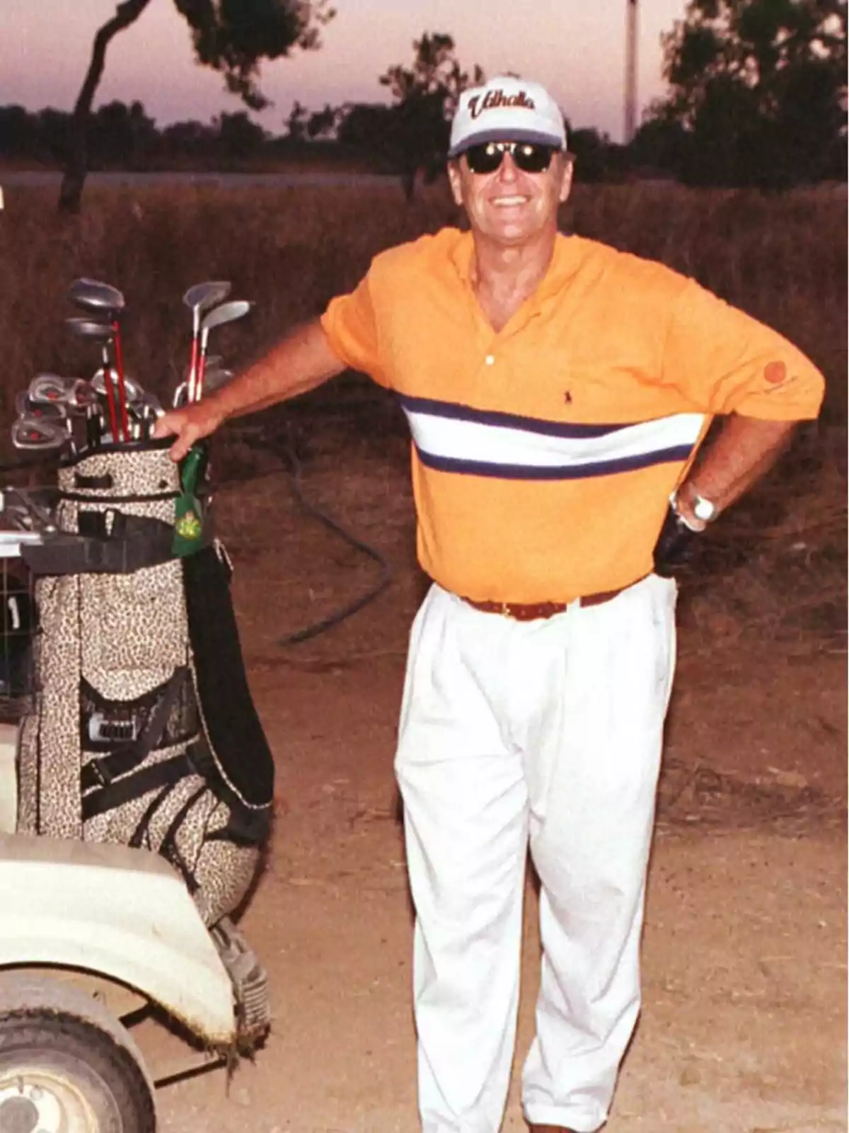 Hombre sonriente con gafas de sol y gorra blanca, vestido con polo naranja y pantalón blanco, apoyado en un carrito de golf junto a una bolsa de palos en un campo al atardecer Hombre sonriente con gafas de sol y gorra blanca, vestido con polo naranja y pantalón blanco, apoyado en un carrito de golf junto a una bolsa de palos en un campo al atardecer