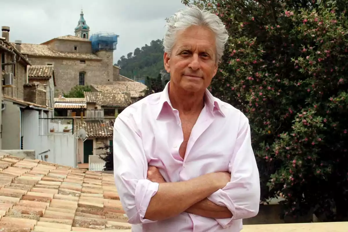 Hombre de cabello canoso con camisa rosa claro de manga larga posando con los brazos cruzados en una terraza con casas de tejas y una iglesia antigua al fondo Hombre de cabello canoso con camisa rosa claro de manga larga posando con los brazos cruzados en una terraza con casas de tejas y una iglesia antigua al fondo