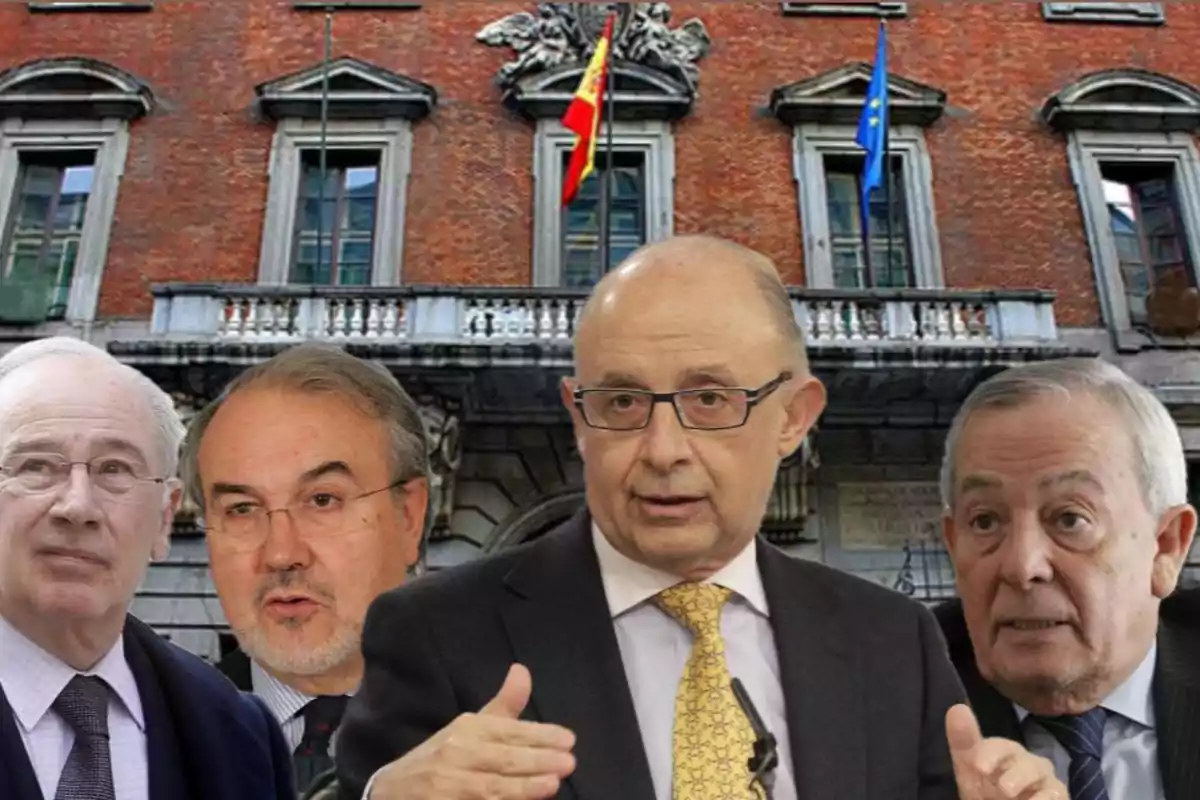 Cuatro hombres mayores con traje y corbata posan frente a un edificio de ladrillo con banderas de España y la Unión Europea.