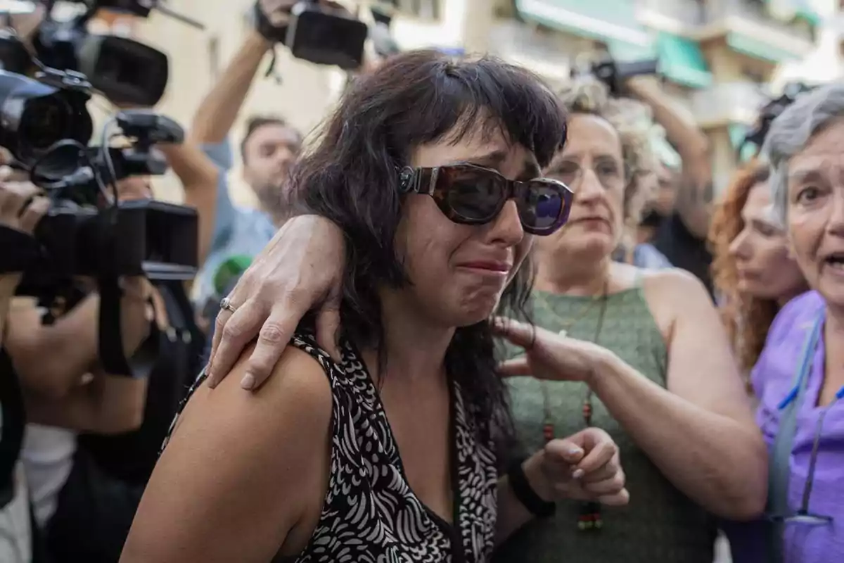 Mujer con gafas de sol llorando mientras es consolada por otras personas en medio de una multitud y cámaras de prensa Mujer con gafas de sol llorando mientras es consolada por otras personas en medio de una multitud y cámaras de prensa