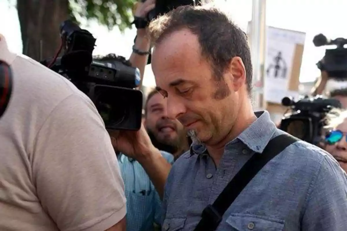 Hombre de mediana edad con expresión seria rodeado de cámaras y periodistas en un evento al aire libre Hombre de mediana edad con expresión seria rodeado de cámaras y periodistas en un evento al aire libre