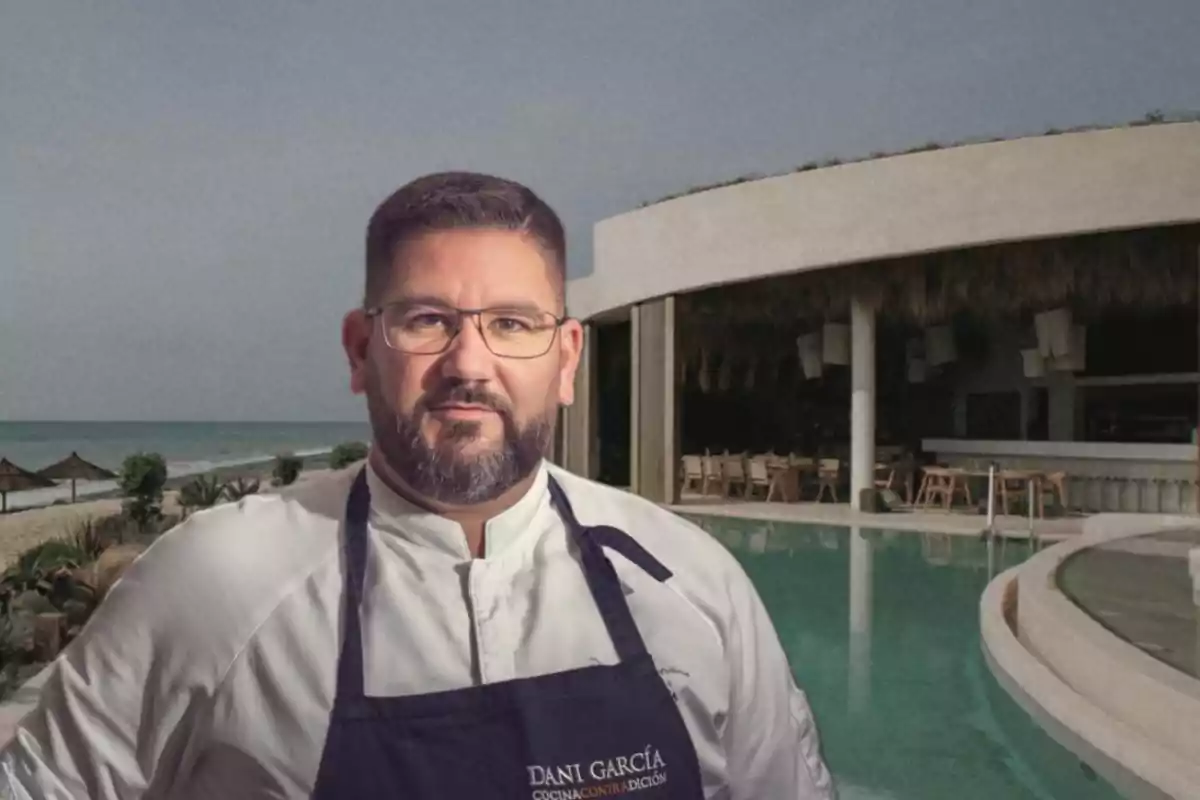 Chef con gafas y barba frente a una piscina y un restaurante junto a la playa