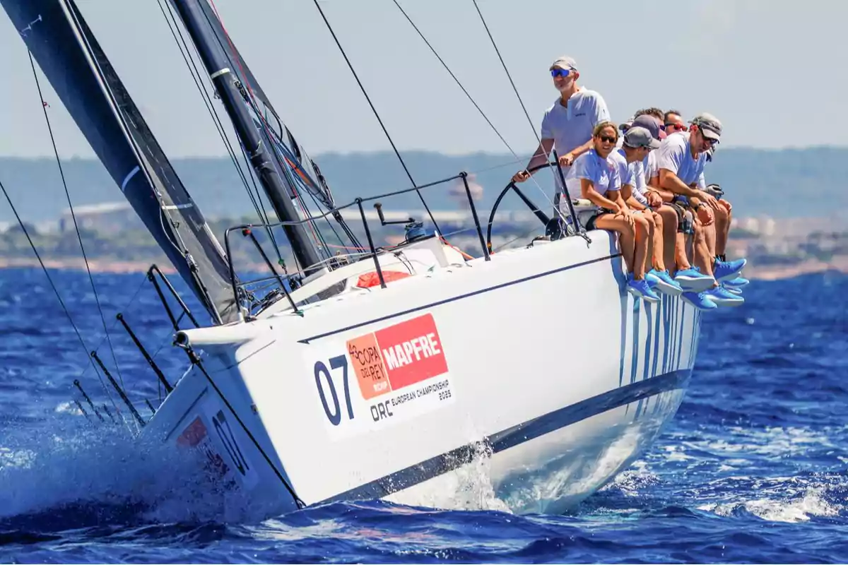Un velero de competición navega a gran velocidad con su tripulación sentada en el borde usando ropa y calzado deportivo mientras el mar está en calma y el cielo despejado Un velero de competición navega a gran velocidad con su tripulación sentada en el borde usando ropa y calzado deportivo mientras el mar está en calma y el cielo despejado