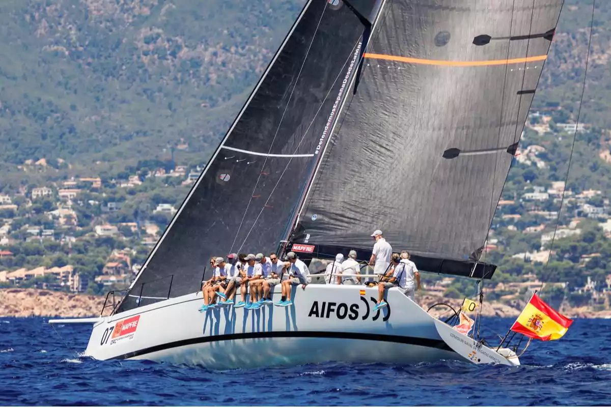 Un velero de competición con varias personas a bordo navega en aguas azules cerca de la costa, con una gran vela negra y una bandera de España ondeando en la parte trasera. Un velero de competición con varias personas a bordo navega en aguas azules cerca de la costa, con una gran vela negra y una bandera de España ondeando en la parte trasera.