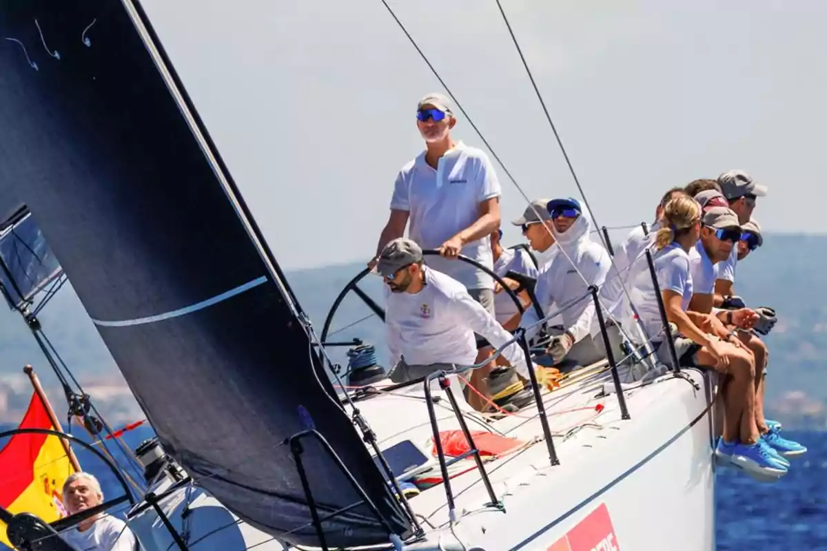 Un grupo de personas navega en un velero durante una competencia en el mar, todos llevan ropa deportiva y gorra, y al fondo se ve una bandera de España. Un grupo de personas navega en un velero durante una competencia en el mar, todos llevan ropa deportiva y gorra, y al fondo se ve una bandera de España.