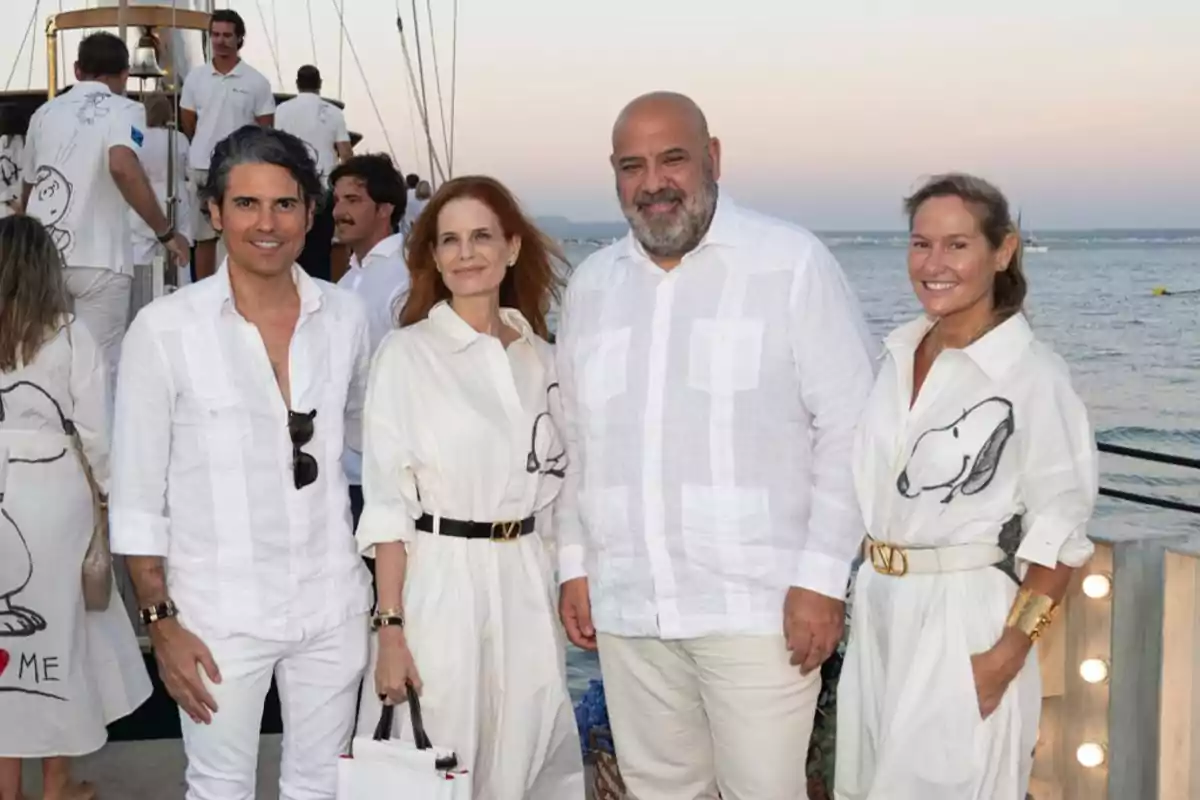 Cuatro personas posan sonrientes junto al mar durante un evento al atardecer, todas vestidas de blanco y dos de ellas llevan prendas con el dibujo de Snoopy.