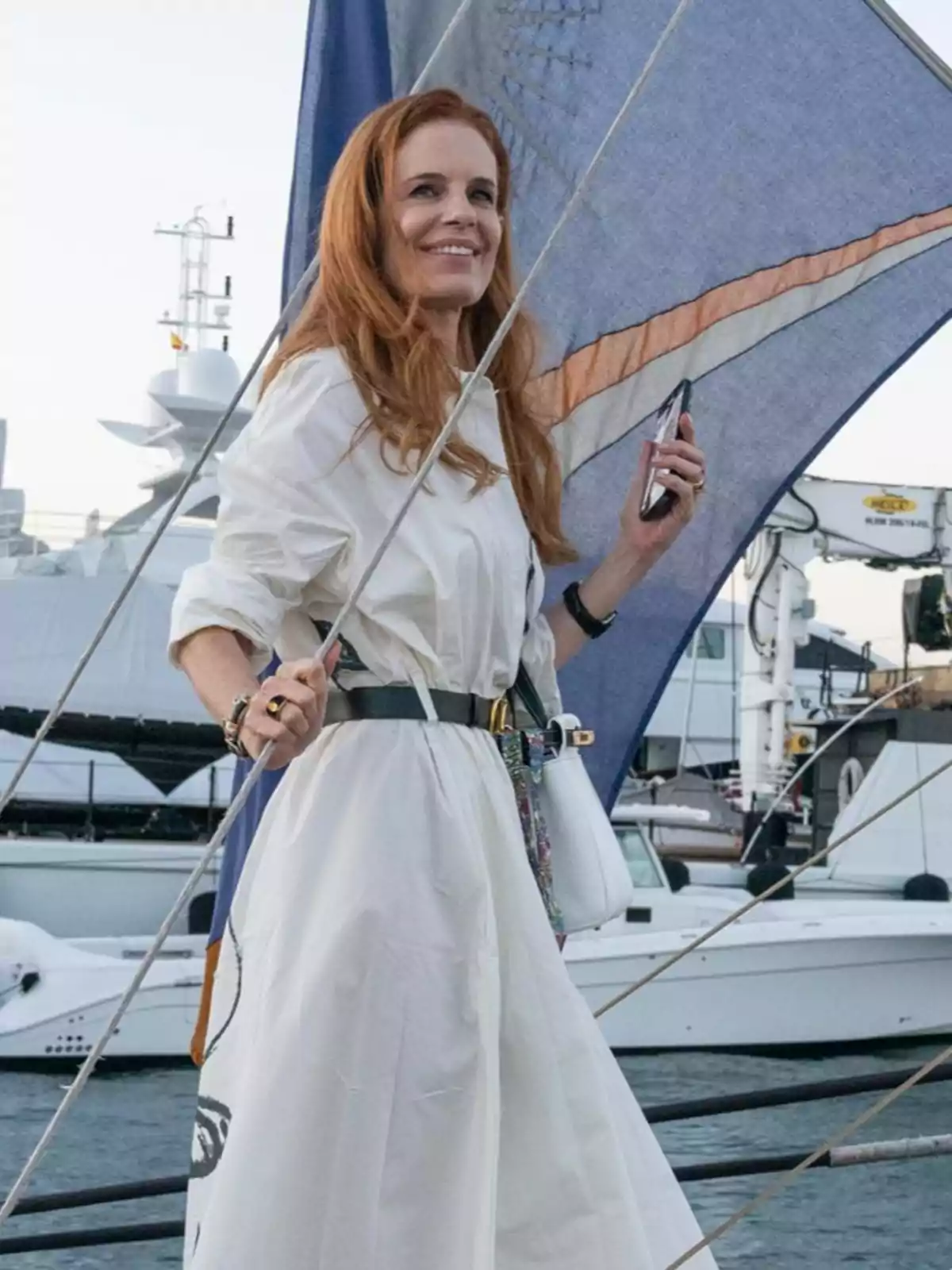 Mujer pelirroja con vestido blanco sonríe mientras sostiene un teléfono móvil a bordo de un velero con yates al fondo Mujer pelirroja con vestido blanco sonríe mientras sostiene un teléfono móvil a bordo de un velero con yates al fondo