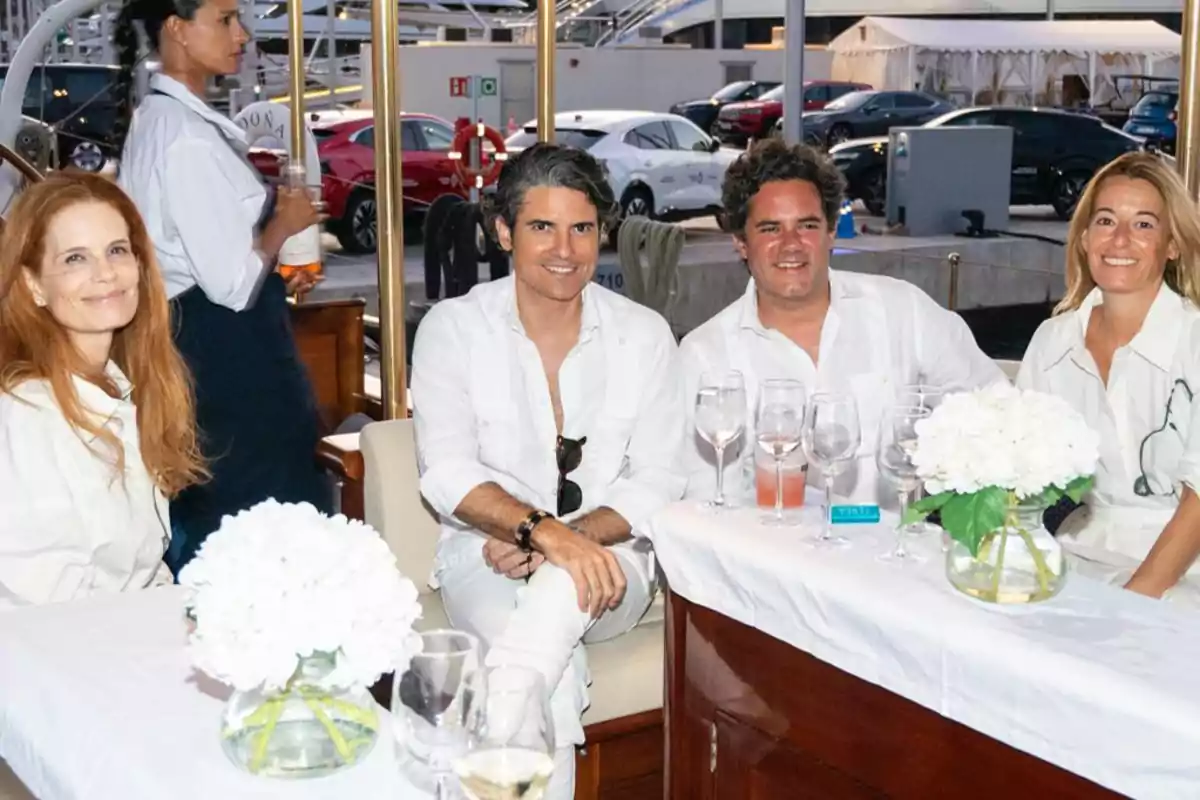 Cuatro personas sonríen sentadas en una mesa decorada con flores blancas y copas de vino, mientras una camarera pasa al fondo en un ambiente elegante al aire libre. Cuatro personas sonríen sentadas en una mesa decorada con flores blancas y copas de vino, mientras una camarera pasa al fondo en un ambiente elegante al aire libre.