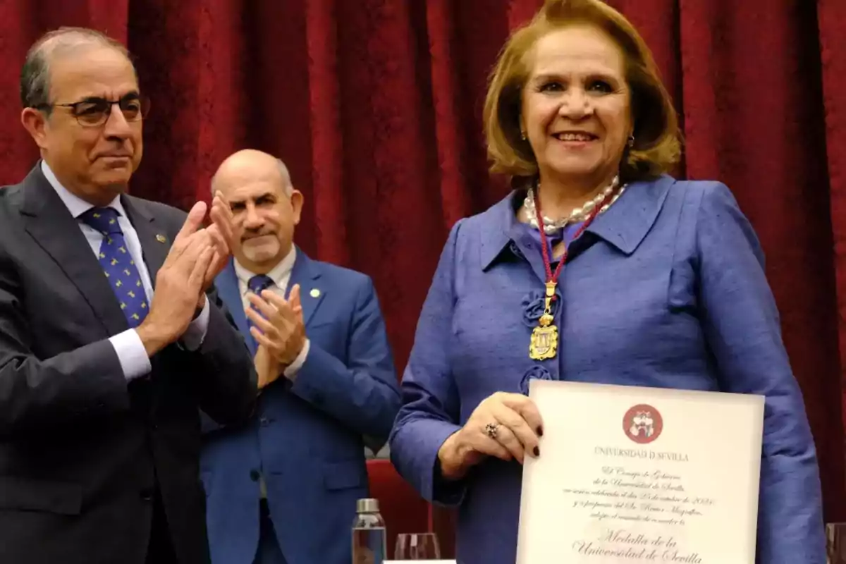 Mujer sonriente sostiene un diploma mientras dos hombres aplauden en un evento formal frente a un fondo de cortinas rojas