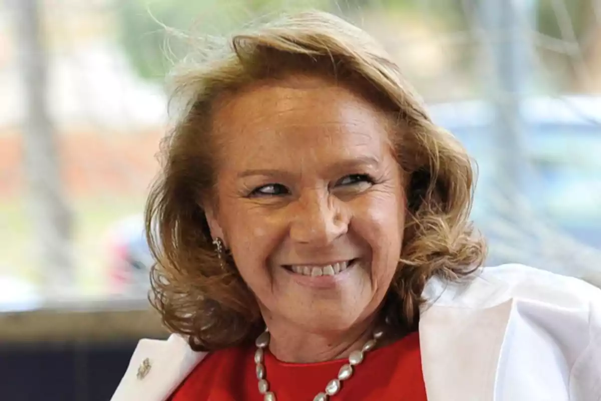 Mujer de cabello rubio sonriente con collar de perlas y chaqueta blanca