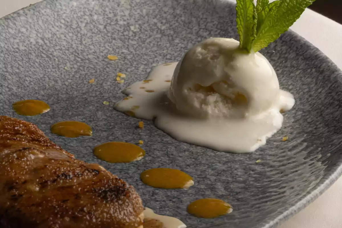Helado de vainilla derritiéndose decorado con una hoja de menta junto a un trozo de postre y gotas de salsa amarilla sobre un plato azul Helado de vainilla derritiéndose decorado con una hoja de menta junto a un trozo de postre y gotas de salsa amarilla sobre un plato azul