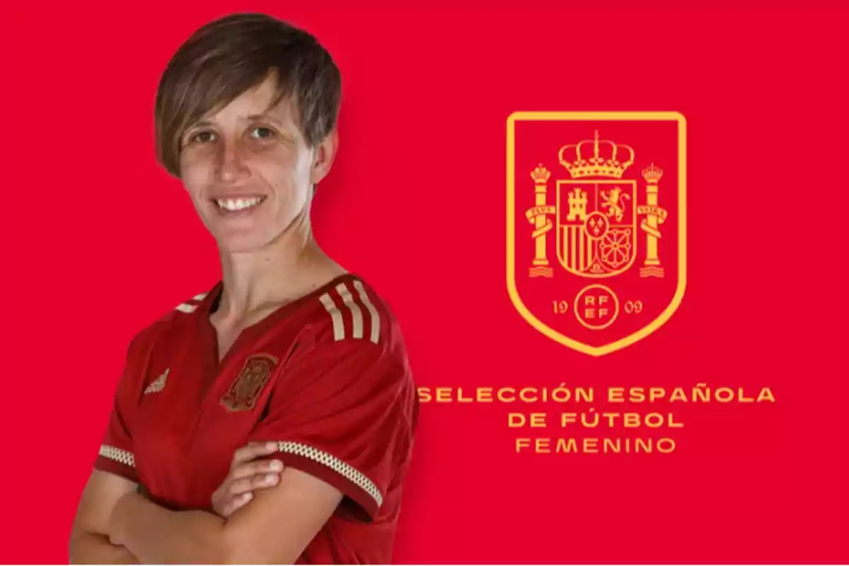 Persona con camiseta roja de la selección española de fútbol femenino posando frente a un fondo rojo con el escudo y el nombre del equipo en amarillo
