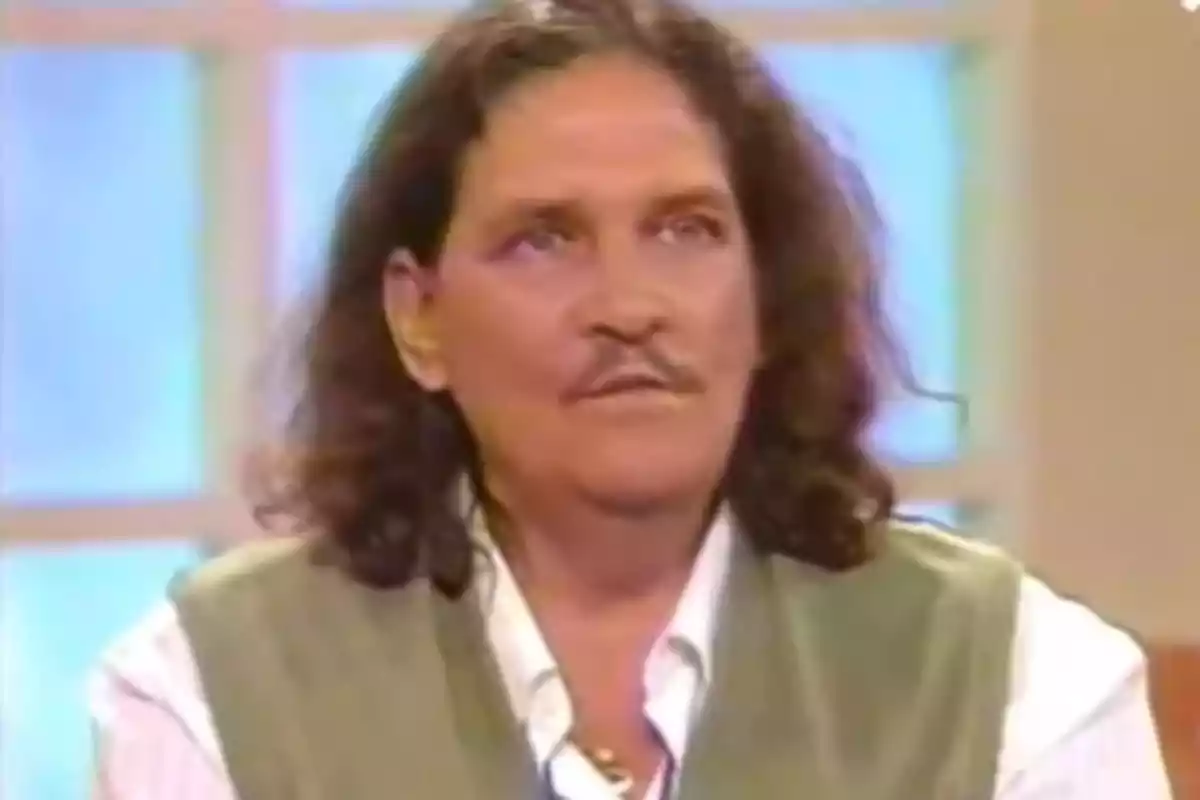 Hombre de cabello largo y bigote vistiendo camisa blanca y chaleco verde sentado en un estudio de televisión Hombre de cabello largo y bigote vistiendo camisa blanca y chaleco verde sentado en un estudio de televisión