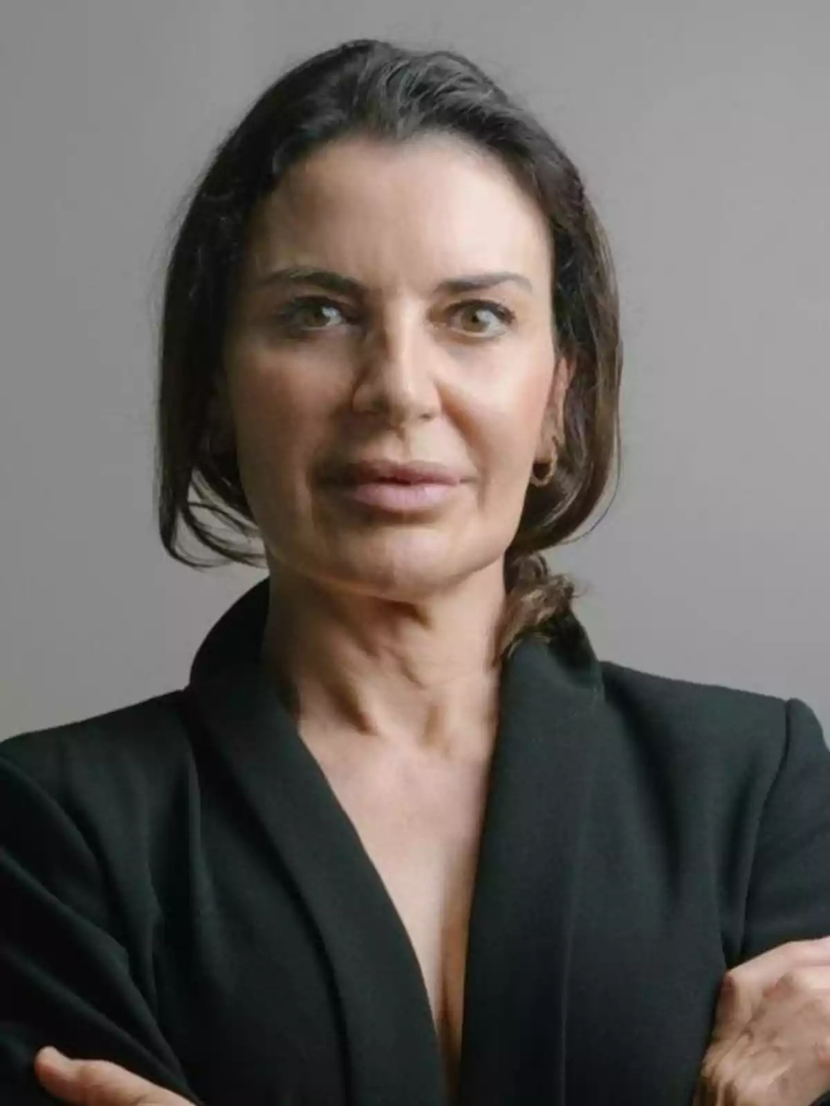 Mujer de cabello oscuro con expresión seria vestida con saco negro y fondo gris Mujer de cabello oscuro con expresión seria vestida con saco negro y fondo gris