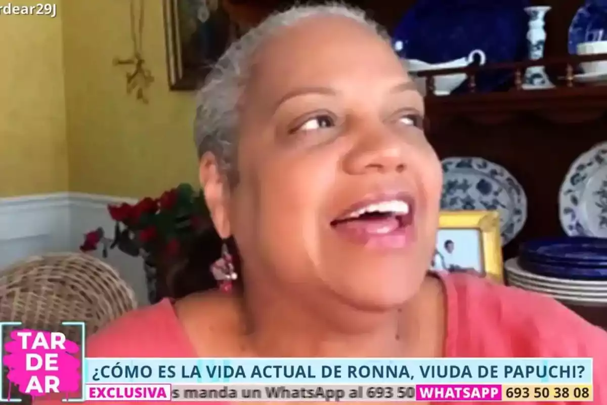 Mujer sonriente de cabello corto y canoso participando en una videollamada, con platos decorativos y flores en el fondo, durante una entrevista televisiva sobre su vida actual Mujer sonriente de cabello corto y canoso participando en una videollamada, con platos decorativos y flores en el fondo, durante una entrevista televisiva sobre su vida actual
