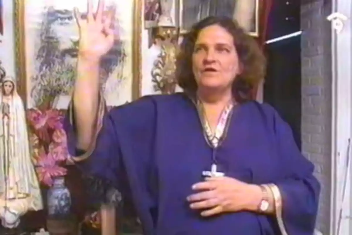 Mujer de cabello rizado y vestido azul levantando una mano con cuatro dedos extendidos en una habitación decorada con imágenes religiosas y flores