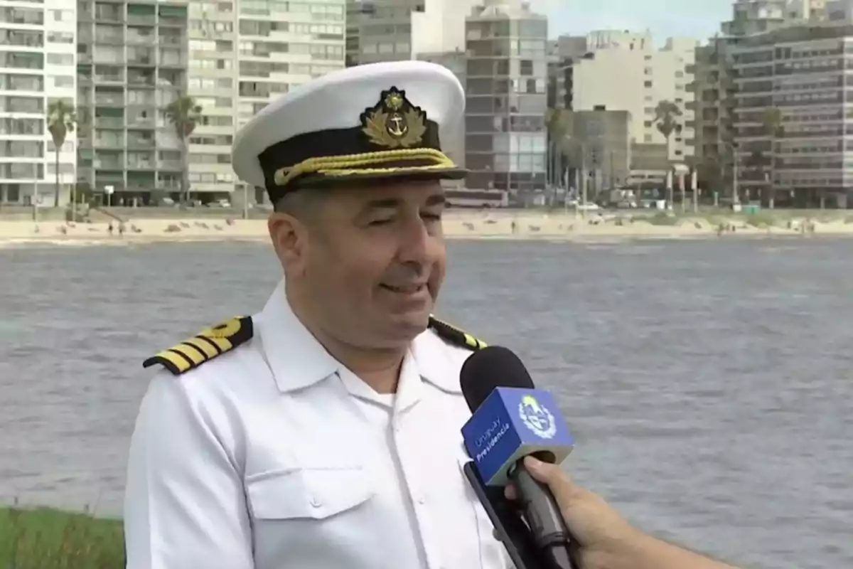 A naval officer in a white uniform is being interviewed by the river with buildings and a beach in the background. A naval officer in a white uniform is being interviewed by the river with buildings and a beach in the background.