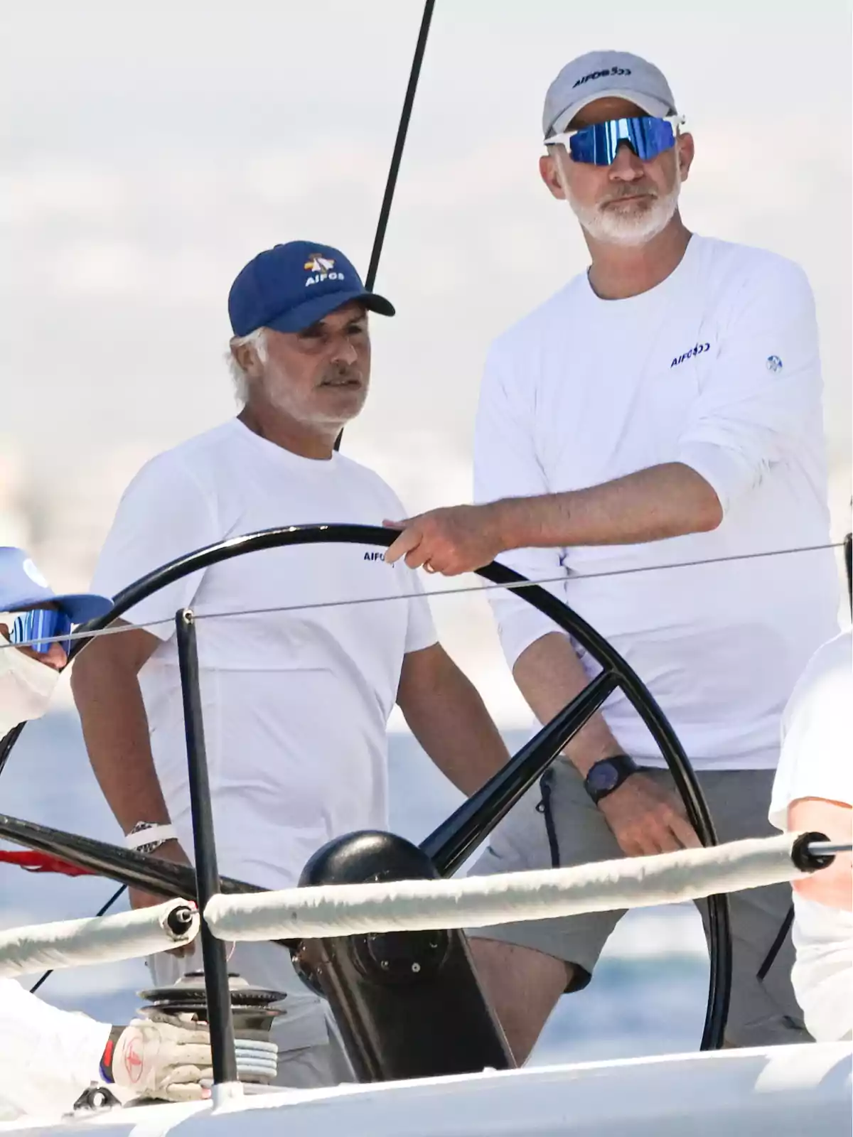 Dos hombres con barba y camisetas blancas navegan en un velero, uno de ellos lleva gafas de sol y gorra, ambos están concentrados en el timón Dos hombres con barba y camisetas blancas navegan en un velero, uno de ellos lleva gafas de sol y gorra, ambos están concentrados en el timón