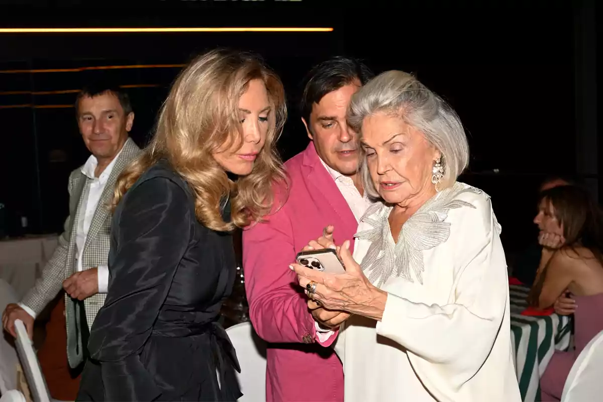Tres personas observan la pantalla de un teléfono móvil en una reunión social mientras otra persona los mira al fondo Tres personas observan la pantalla de un teléfono móvil en una reunión social mientras otra persona los mira al fondo
