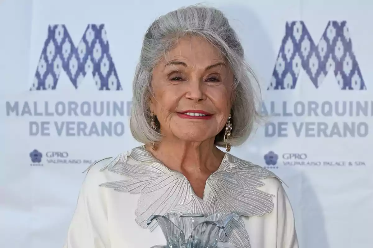 Mujer mayor de cabello canoso y corto sonríe mientras sostiene un trofeo de cristal en un evento con fondo que dice Mallorquines de Verano