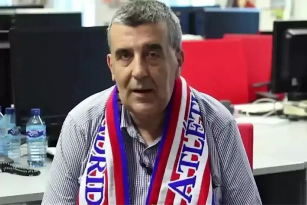 Hombre de cabello canoso con bufanda del Atlético de Madrid sentado en una oficina con botellas de agua y computadoras al fondo Hombre de cabello canoso con bufanda del Atlético de Madrid sentado en una oficina con botellas de agua y computadoras al fondo
