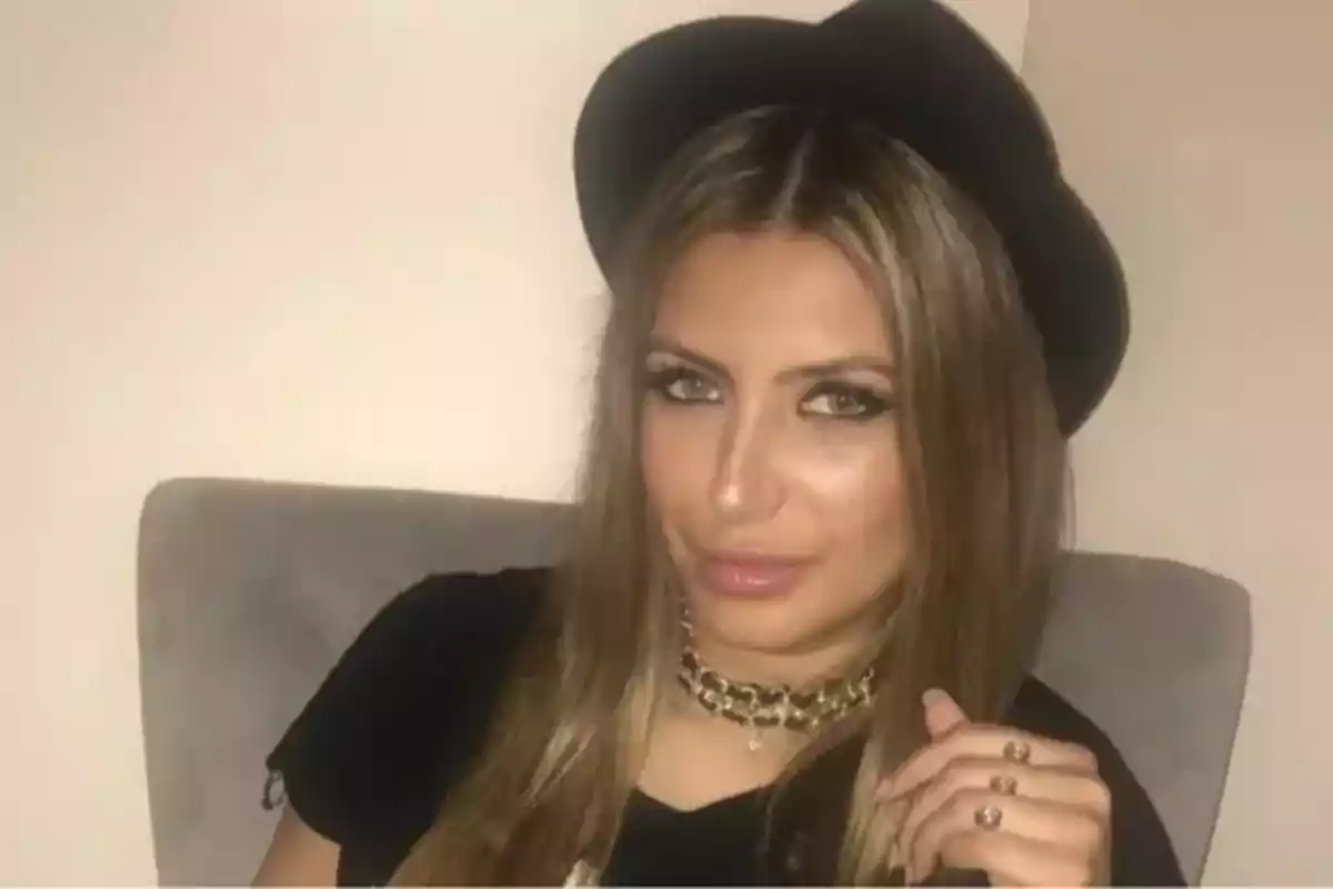 Mujer de cabello largo y rubio sentada en una silla, usando un sombrero negro y un collar dorado, con maquillaje marcado y expresión seria. Mujer de cabello largo y rubio sentada en una silla, usando un sombrero negro y un collar dorado, con maquillaje marcado y expresión seria.