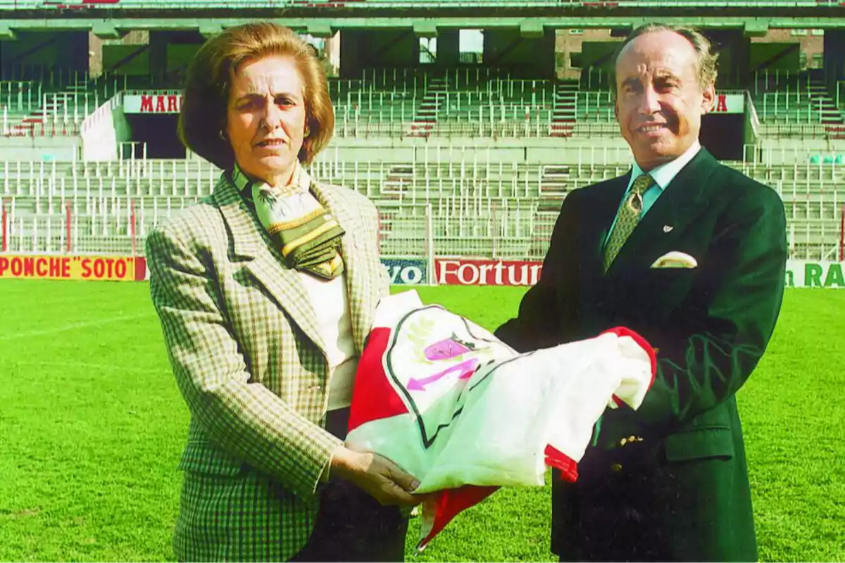 Dos personas elegantemente vestidas sostienen una bandera en el centro de un estadio de fútbol vacío Dos personas elegantemente vestidas sostienen una bandera en el centro de un estadio de fútbol vacío