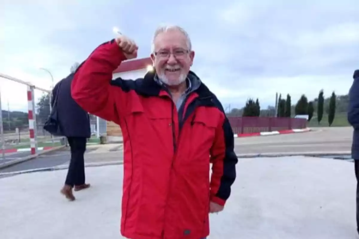 Hombre mayor con barba y gafas sonríe mientras levanta el brazo en señal de fuerza, lleva una chaqueta roja y está al aire libre Hombre mayor con barba y gafas sonríe mientras levanta el brazo en señal de fuerza, lleva una chaqueta roja y está al aire libre