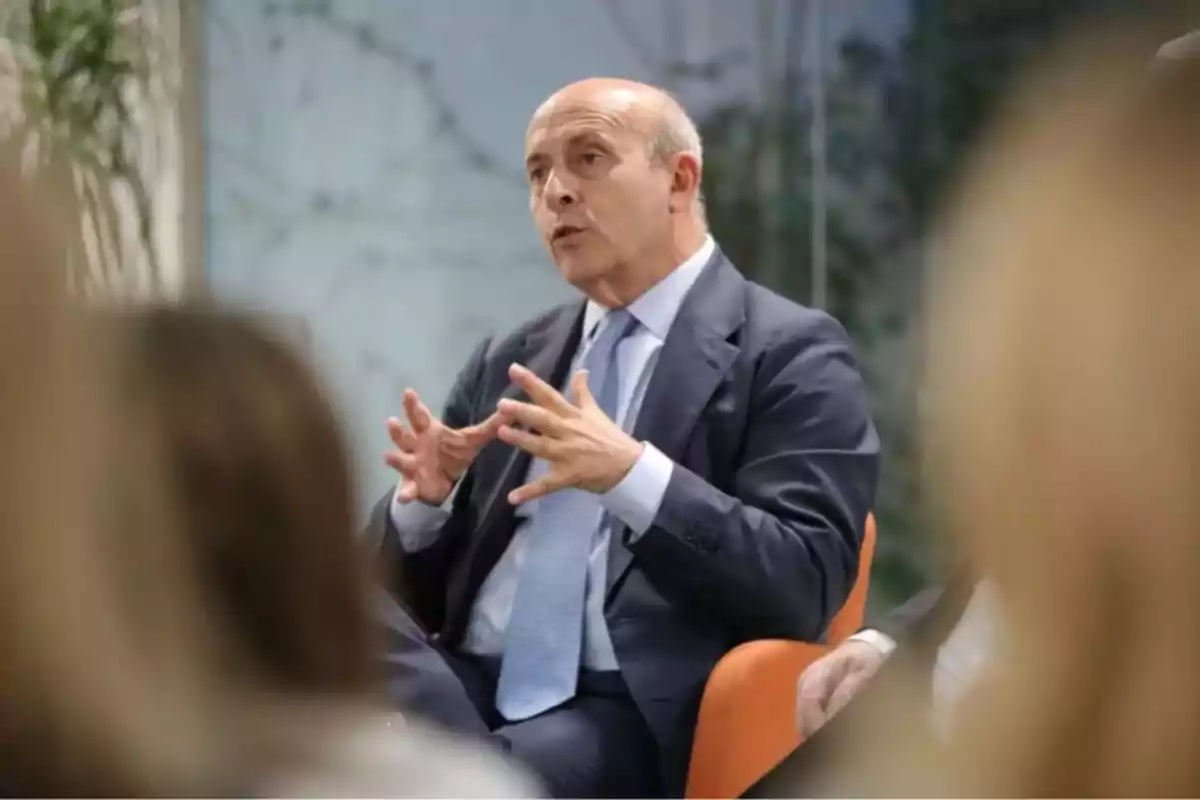 Hombre calvo con traje y corbata azul claro sentado en una silla naranja gesticulando con las manos mientras habla en un entorno profesional Hombre calvo con traje y corbata azul claro sentado en una silla naranja gesticulando con las manos mientras habla en un entorno profesional