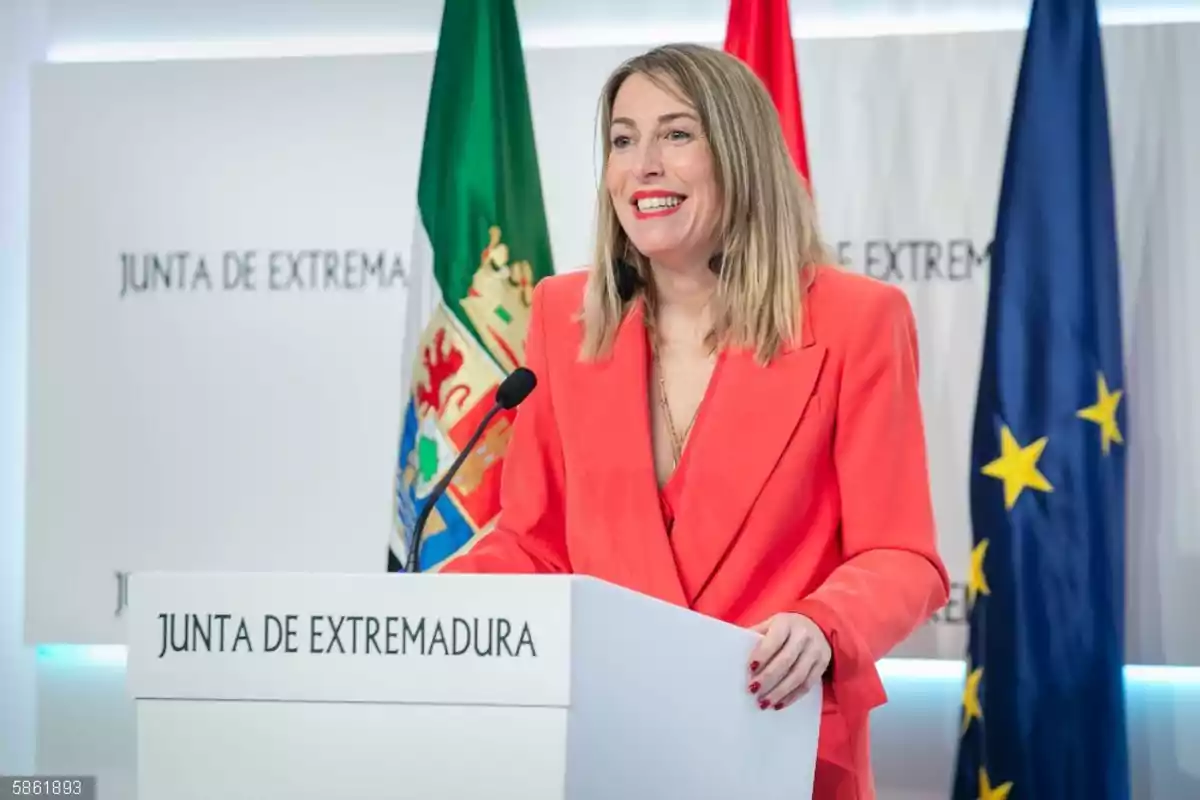 Mujer sonriente con chaqueta roja hablando en un atril de la Junta de Extremadura con banderas de fondo Mujer sonriente con chaqueta roja hablando en un atril de la Junta de Extremadura con banderas de fondo