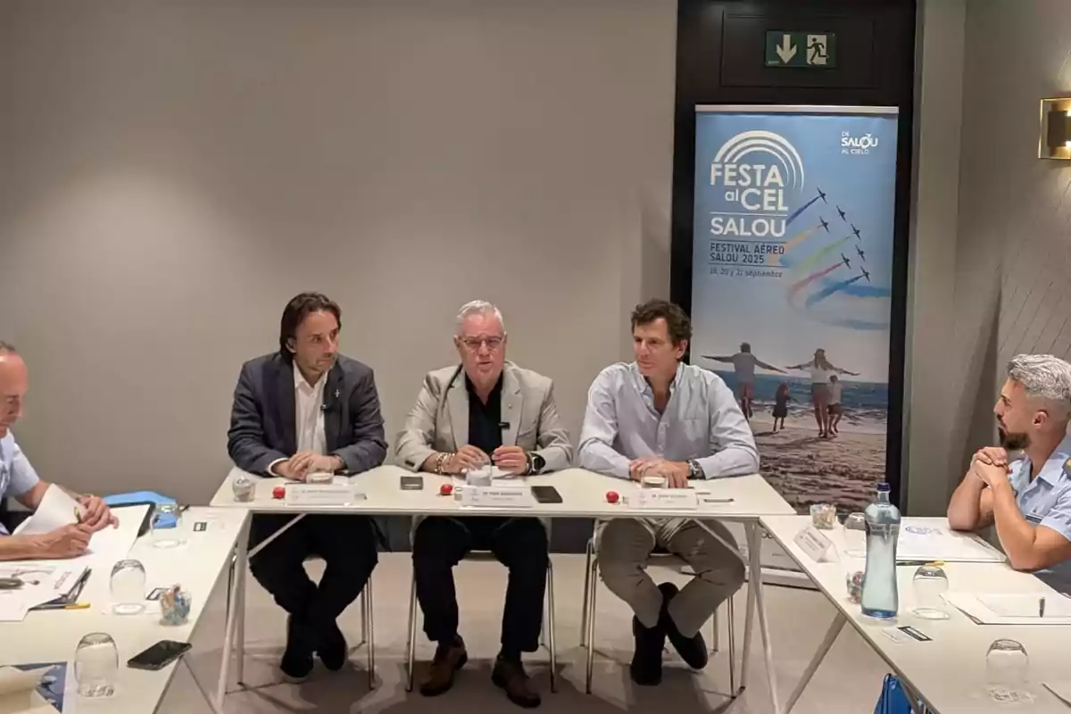 Cinco hombres sentados en una mesa de reunión con papeles y botellas de agua, al fondo un cartel promocional del festival aéreo Festa al Cel Salou
