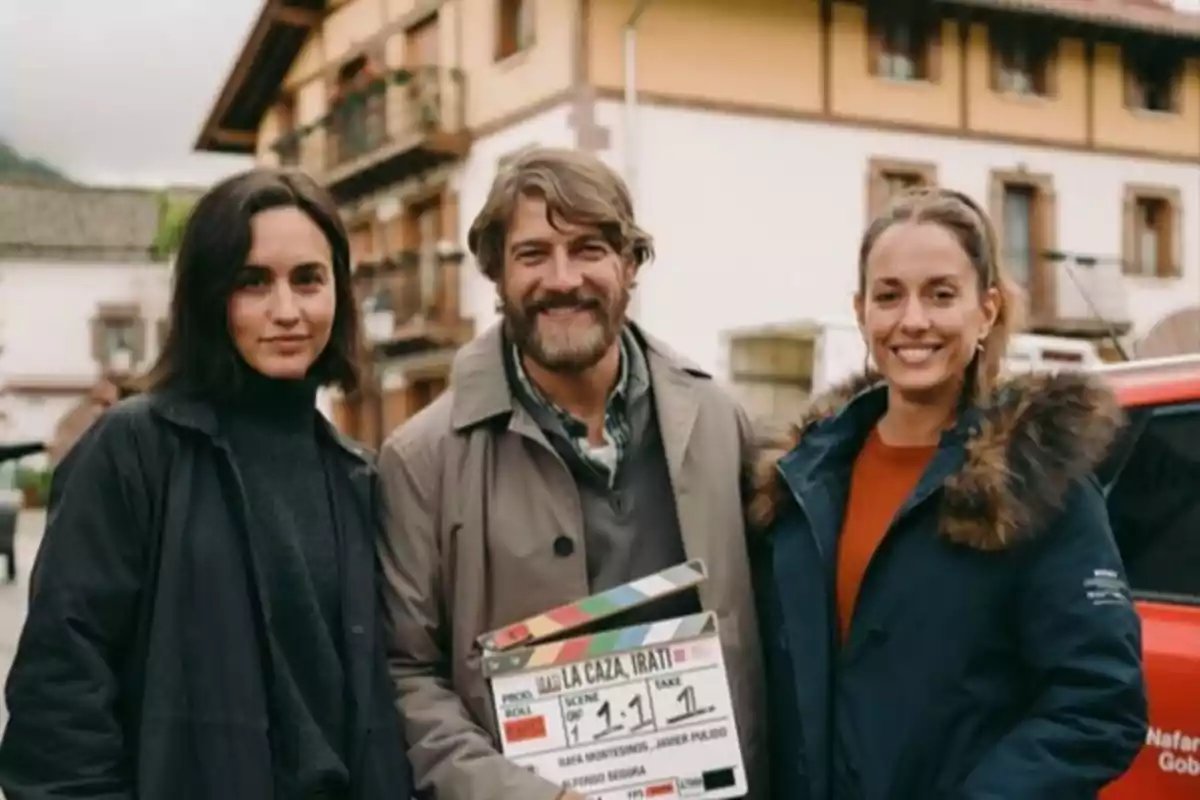 Tres personas sonríen a la cámara en una calle, una de ellas sostiene una claqueta de cine Tres personas sonríen a la cámara en una calle, una de ellas sostiene una claqueta de cine