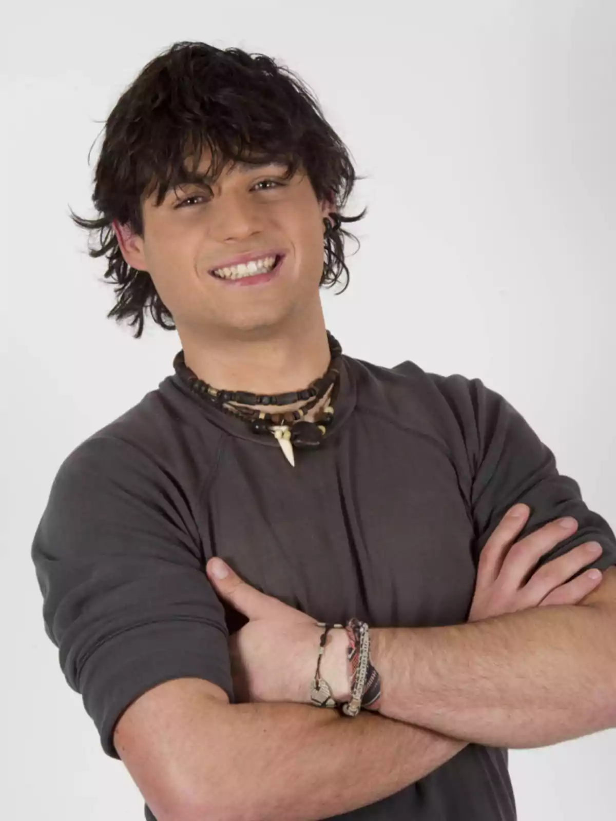 Joven de cabello oscuro y ondulado sonriendo con los brazos cruzados, lleva una camiseta gris y varios collares y pulseras Joven de cabello oscuro y ondulado sonriendo con los brazos cruzados, lleva una camiseta gris y varios collares y pulseras