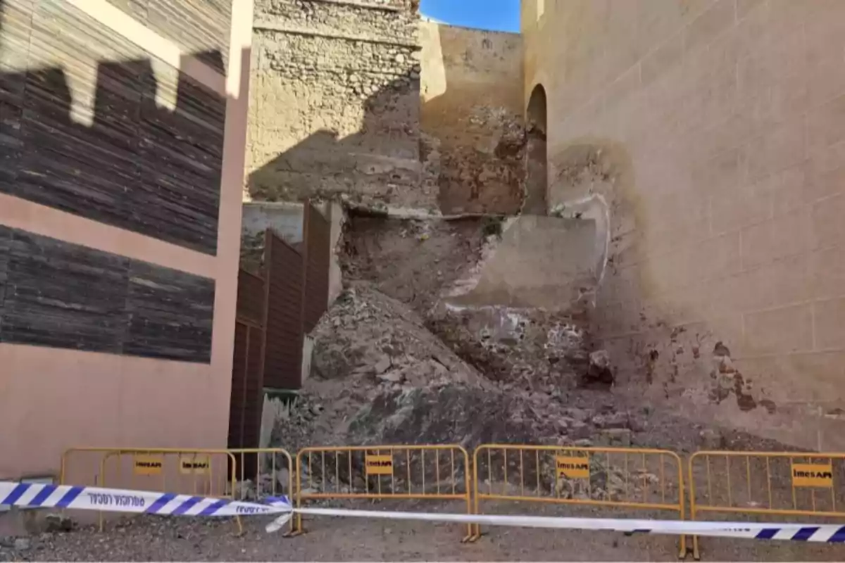 Derrumbe de un muro antiguo con escombros en el suelo y una zona acordonada con vallas amarillas y cinta de seguridad