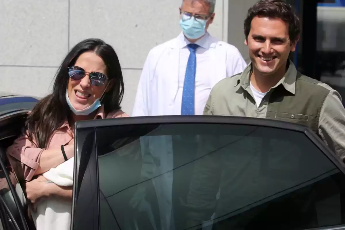 Una mujer con gafas de sol y mascarilla bajada sonríe junto a un hombre mientras salen de un edificio, con un médico al fondo y una puerta de coche en primer plano Una mujer con gafas de sol y mascarilla bajada sonríe junto a un hombre mientras salen de un edificio, con un médico al fondo y una puerta de coche en primer plano