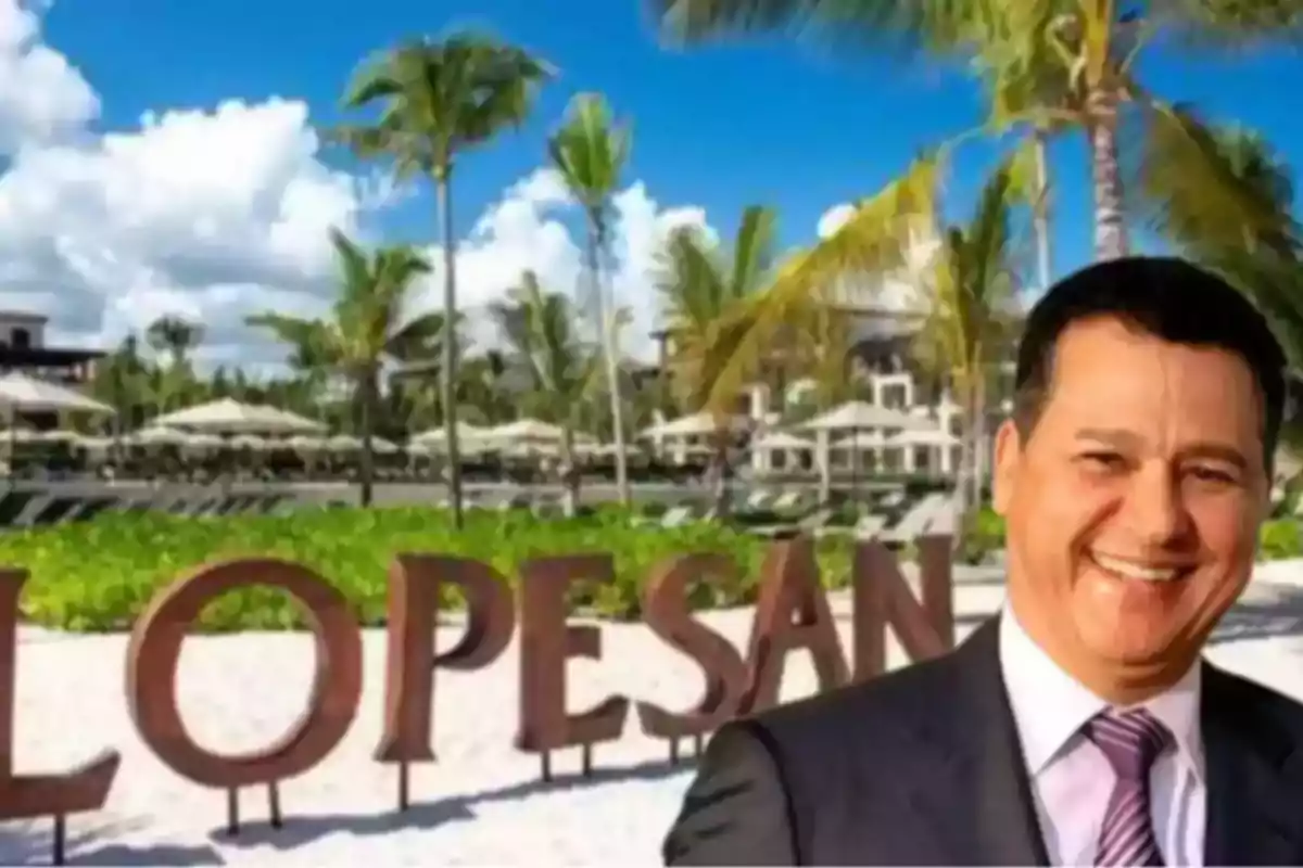 Hombre sonriente con traje frente a un letrero grande de Lopesan en una playa con palmeras y sombrillas Hombre sonriente con traje frente a un letrero grande de Lopesan en una playa con palmeras y sombrillas