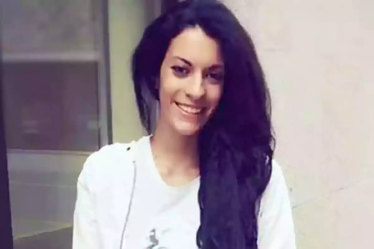 Mujer joven de cabello largo y oscuro sonriendo frente a una pared clara Mujer joven de cabello largo y oscuro sonriendo frente a una pared clara