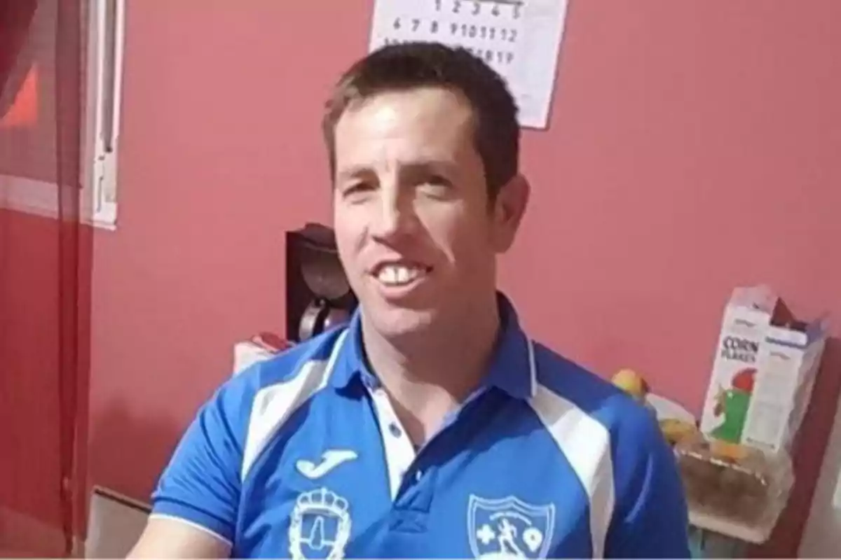 Hombre sonriente con camiseta azul deportiva en una cocina con paredes rosadas y una caja de cereal al fondo