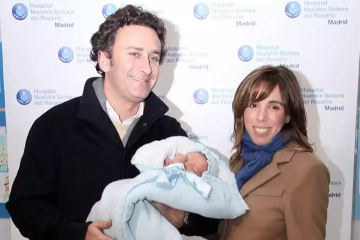 Una pareja sonriente sostiene a un bebé envuelto en una manta azul frente a un fondo con el logo del Hospital Nuestra Señora del Rosario en Madrid Una pareja sonriente sostiene a un bebé envuelto en una manta azul frente a un fondo con el logo del Hospital Nuestra Señora del Rosario en Madrid