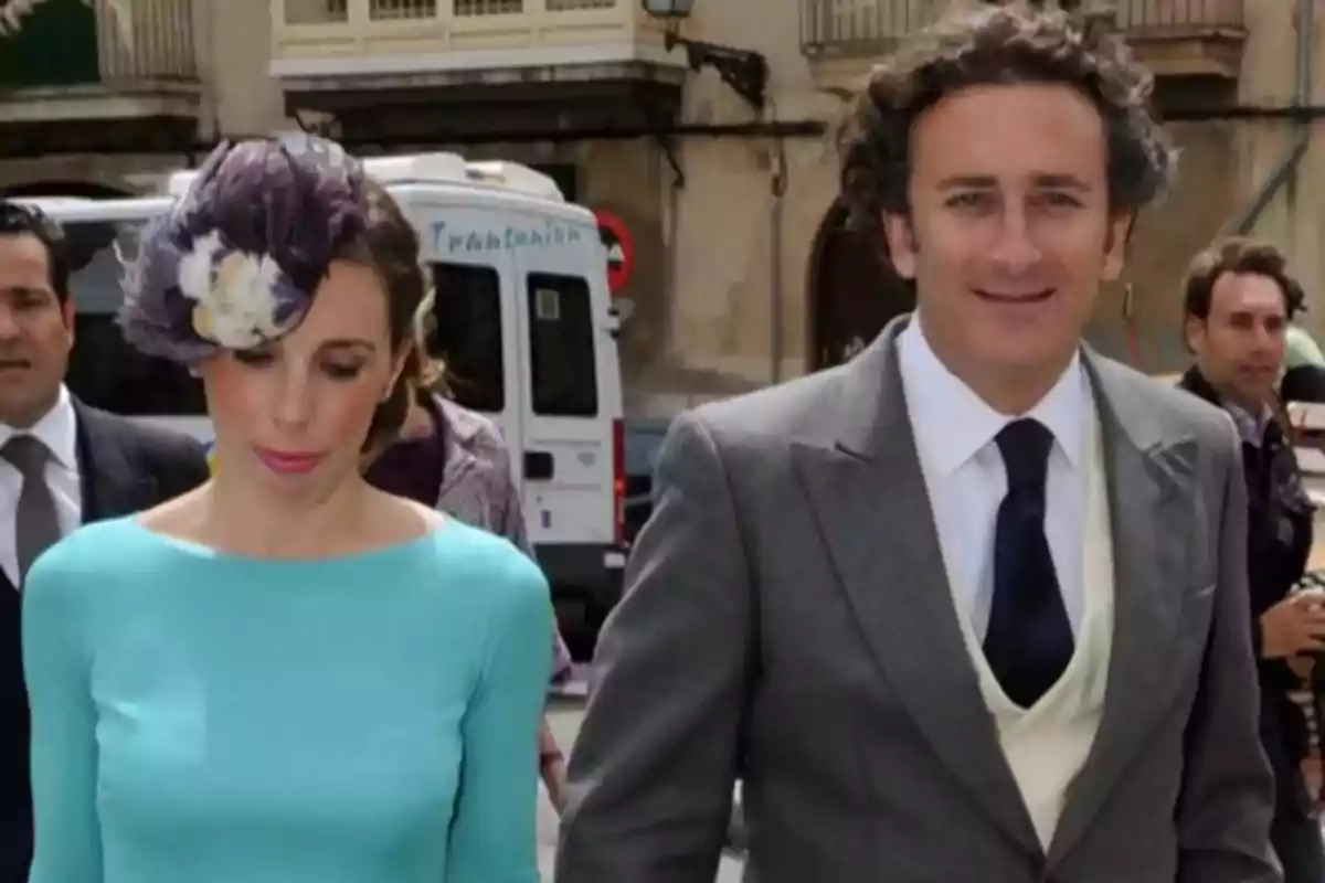 Pareja elegantemente vestida caminando por la calle, la mujer lleva un vestido azul claro y un tocado con plumas, el hombre lleva un traje gris con chaleco claro y corbata oscura Pareja elegantemente vestida caminando por la calle, la mujer lleva un vestido azul claro y un tocado con plumas, el hombre lleva un traje gris con chaleco claro y corbata oscura